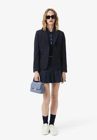 Donna che indossa un blazer blu navy, una gonna plissettata e una polo, con una piccola borsa blu, occhiali da sole neri e sneakers bianche.