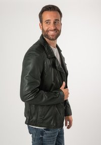 Giacca da moto nera in pelle con spalline, tasche con zip e design slim fit. Indossata sopra una camicia bianca e abbinata a jeans blu.