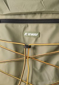 Λαδί ύφασμα με μαύρο φερμουάρ, κίτρινο-μαύρο πλεγμένο κορδόνι και πράσινη ορθογώνια ετικέτα με την επιγραφή "K-WAY" ραμμένη στο ύφασμα.