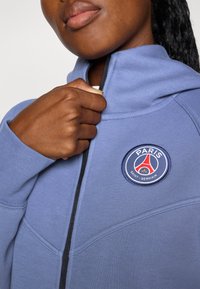 Blå hettegenser med glidelås i kragen, teksturert stoff, med et sirkulært Paris Saint-Germain-logo på brystet. Svart glidelåsdetalj.
