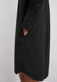 Zwarte shirtjurk met lange mouwen en knoopmanchetten, voorzien van zijzakken en een split aan de zijkant van de zoom voor meer bewegingsruimte.