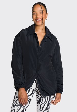 Schwarze, oversized Jacke mit Knopf-Front, weitem Kragen und elastischen Bündchen. Getragen über weißen Leggings mit einem schwarzen Wirbelmuster.