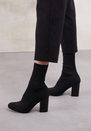 Botines de tacón negro con un upper tipo calcetín acanalado, superficie lisa, punta redonda y un tacón de bloque para mayor estabilidad.