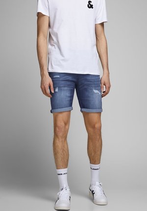 Denim shorts i mellemblå med en slidt finish, opfoldede manchetter og en tætsiddende design, parret med en ensfarvet hvid t-shirt og hvide sneakers.