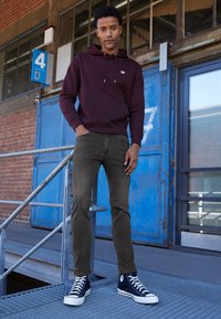 Junger Mann steht auf metallenen Stufen, eine Hand in der Tasche, trägt einen weinroten Kapuzenpullover, dunkle Jeans und navyblaue High-Top-Sneaker neben einer blauen Industrierampe.