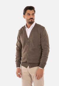 Cardigan marrone lavorato a maglia con scollo a V, caratterizzato da sei bottoni frontali, polsini a costine e orlo aderente, indossato sopra una camicia bianca.