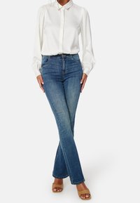 Chemise blanche à manches longues avec col, associée à un jean évasé en denim bleu et des sandales compensées beiges. Texture lisse, style ajusté.