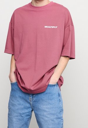 Luźna fioletowa koszulka z białym napisem "MULTIPLY" na przodzie; wykonana z miękkiej bawełny, z krótkimi rękawami i podwiniętym dekoltem.