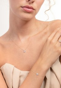 Goldkette und Armband mit einem kleinen, sternförmigen Kristallakzent. Weich drapierter Stoff in neutralem Farbton. Sichtbare, glatte Hautstruktur.