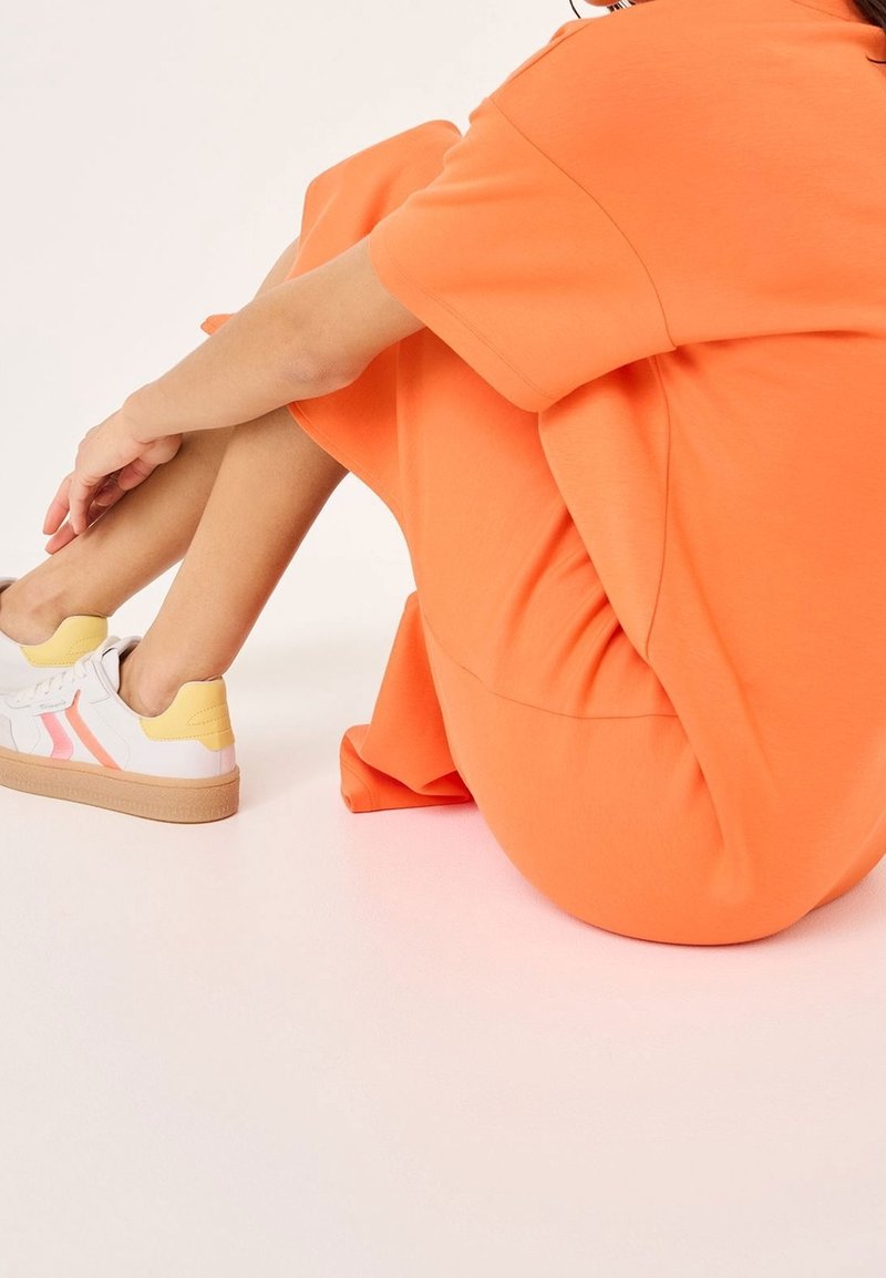 Personne assise en tailleur portant une robe orange et des baskets blanches avec des accents roses et jaunes sur un fond blanc.