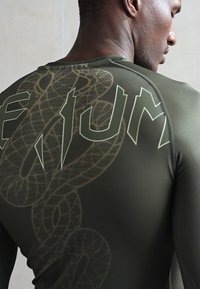 Uomo che indossa una camicia a maniche lunghe verde oliva con una grande grafica di serpente e testo in grassetto sulla schiena, visto di spalle e di lato.