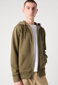 Olijfgroene zip-up hoodie van zachte stof met een kangoeroezak, een trekkoord capuchon en ribboord aan de mouwen en onderkant. Draag het over een wit T-shirt.