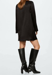 Schwarzes Satin-Kleid mit langen Ärmeln und lockerer Passform, mit einem geschwungenen Saum. Kombiniert mit knielangen schwarzen Lederstiefeln mit einem blockigen Absatz.