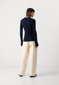 Haut à manches longues côtelé marine au design ajusté, associé à un pantalon large crème, vu de dos sur un fond uni.