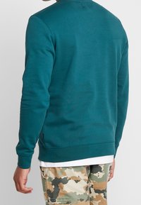 Sudadera en color verde azulado con puños y dobladillo acanalados. Tejido suave y texturizado. Llevada sobre una camisa blanca, combinada con pantalones de camuflaje.
