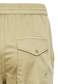 Gros plan sur un short cargo beige montrant une ceinture élastique et une poche à rabat avec un bouton sur le côté.