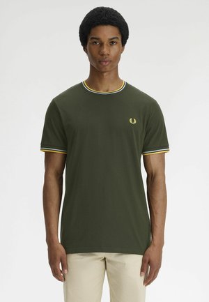 Joven de pie mirando al frente, lleva una camiseta verde oliva con ribetes amarillos y blancos en el cuello y las mangas, y pantalones beige.