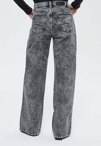 Personne portant un jean gris à taille haute et jambes larges avec des poches arrière et des chaussures noires, se tenant contre un fond uniforme.