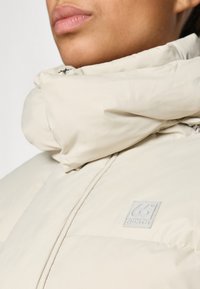 Licht beige pufferjack met een hoge kraag en een opvallende logopatch op de borst. Gladde, glanzende textuur met ritssluiting.