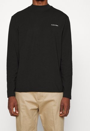 Homme portant une chemise noire à manches longues Calvin Klein avec un pantalon beige, debout devant un fond blanc uni.