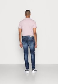 Světlounce růžové tričko, úzké modré džíny s bledými detaily a bílé tenisky s černými akcenty, pohled zezadu na jednobarevném pozadí.