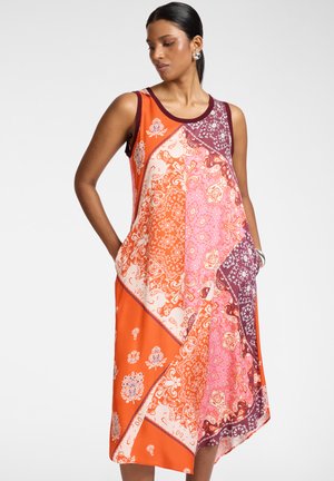 Donna con capelli scuri che indossa un vestito patchwork senza maniche multicolore in arancione, rosa e bordeaux con motivi floreali e paisley, mani nelle tasche.