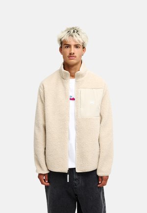 Jonge man met gebleekt haar draagt een beige fleecejack met een borstzak over een wit overhemd en een donkere broek, staand tegen een effen achtergrond.