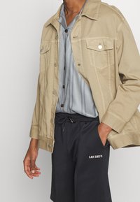 Chaqueta de mezclilla beige sobre una camisa de botones a rayas azul claro, combinada con pantalones cortos negros que llevan el texto "LES DEUX" en blanco.