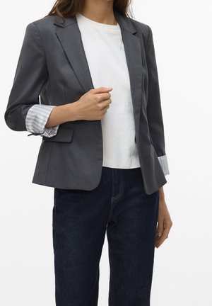 Femme portant un blazer gris foncé avec des poignets rayés retroussés sur une chemise blanche et un jean bleu foncé, ajustant le devant du blazer de la main.