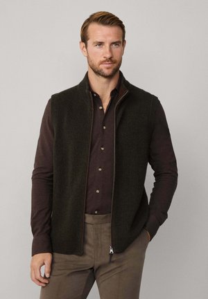 Dunkelgraue Strickweste mit gerippter Textur, mit Reißverschluss vorne und hohem Kragen, getragen über einem dunkelblauen Hemd mit Knöpfen. Das Model steht mit den Händen in den Taschen.