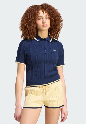 Joven con cabello rizado que lleva una camiseta polo de punto azul marino y pantalones cortos de punto color crema con ribete azul marino, de pie frente a un fondo liso.
