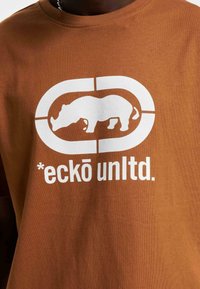 Brun bomulds t-shirt med et hvidt grafisk motiv af et næsehorn inden i en cirkulær ramme og teksten “*eckō unltd.” nedenunder.
