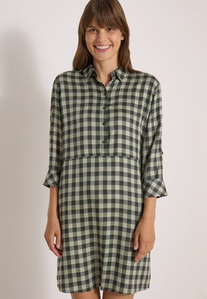 Femme souriante portant une robe chemise à carreaux verts et gris avec des manches trois-quarts sur un fond clair uni.