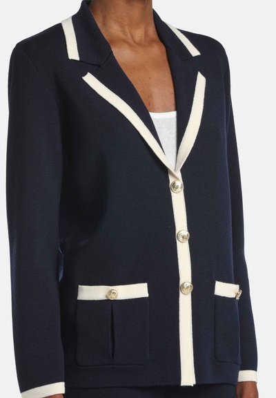 Navy cardigan with cream trim, featuring a notched collar, gold buttons, and front pockets. Made of textured knit fabric.
