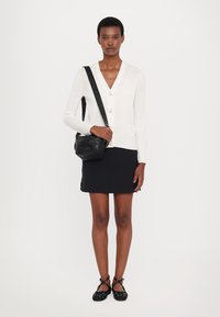 Bílý kardigan s dlouhým rukávem a knoflíky, zkombinovaný s černou mini sukni a černými plochými botami s překříženými pásky. Černá crossbody taška.