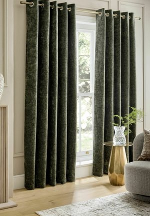 Dunkelgrüne Samtvorhänge hängen an einer goldenen Stange in einem Wohnzimmer mit einer Glasvase auf einem goldenen Beistelltisch und einem grauen Sofa.