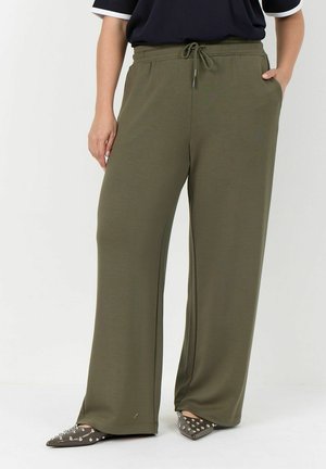 Persoon draagt olijfgroene wijde broek met elastische tailleband en trekkoord, zwart topje en puntige schoenen met metalen studs.