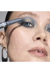 Eyeliner grigio applicato vicino agli occhi blu, con ombretti metallici argento e blu sulle palpebre, in primo piano su pelle liscia e labbra lucide.