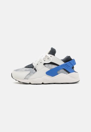 Baskets de sport blanches et grises avec des accents noirs, des lacets blancs et une sangle bleue proéminente au talon, vues de côté sur un fond uni.