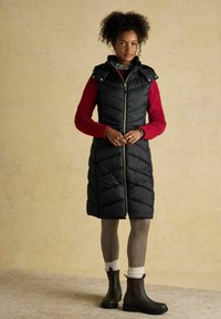 Czarna pikowana kamizelka puchowa z zapięciem na zamek, wysokim kołnierzem i napami na ramionach. Czerwona sweter z dzianiny pod spodem, beżowe legginsy w wzory i brązowe botki za kostkę.