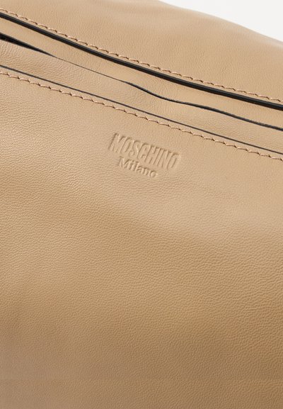 Sac à main en cuir beige avec une texture lisse, des coutures discrètes et un détail de rayure noire. Logo embossé portant la mention "MOSCHINO Milano."