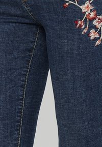 Jeans de denim azul oscuro con bordado floral en rosa y blanco en la parte frontal. La tela tiene una textura suave y un diseño de corte ajustado.