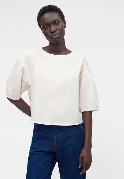 Γυναίκα με κοντά κατσαρά μαλλιά, φορώντας λευκό μπλουζάκι με φουσκωτά μανίκια και σκούρο μπλε τζιν με ψηλή μέση, όρθια σε απλό φόντο.