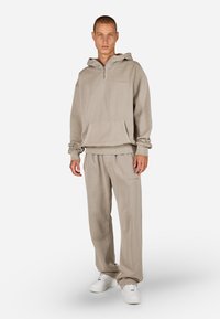 Bežová mikina s kapucňou a polovičným zipsom, predná vrecko a manžety na rukávoch, skombinovaná s voľnými nohavicami v rovnakom štýle, nosená s bielymi teniskami.