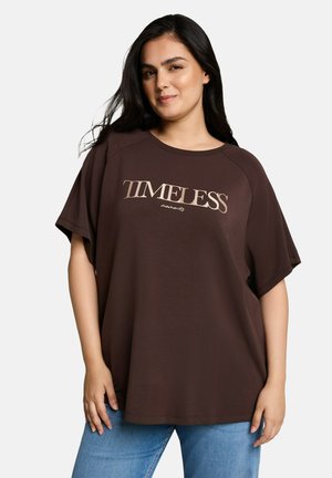 Kobieta z długimi ciemnymi włosami, ubrana w luźny brązowy t-shirt z tekstem "TIMELESS moments" i niebieskie dżinsy na jednolitym tle.