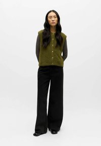Vestenka z texturovaného pleteného materiálu v olivově zelené barvě s knoflíky, vrstvená přes šedou košili s dlouhým rukávem; spojena s širokými černými kalhotami a lesklými botami.