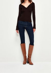 Haut marron à manches longues côtelées avec un col en V, associé à un jean skinny bleu foncé et des bottes en cuir marron à talons montant jusqu'aux genoux.