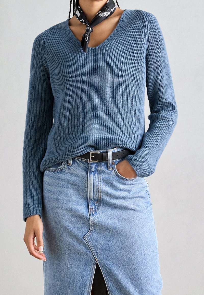 Personne portant un pull en maille côtelée bleu, une jupe en denim bleu clair avec une fente à l'avant, une ceinture noire et un foulard noir à motifs autour du cou.
