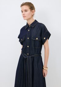 Denim-mekko, tummansininen kultaisilla napilla, lyhyet hihat, kaksi etutaskua, vyötärövyö ja kontrastiväriset ompelutiedot.