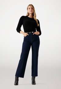 Maglione nero a maglia con maniche lunghe, abbinato a jeans a gamba larga di colore blu scuro a vita alta. Cintura nera con fibbia in metallo, stivaletti.